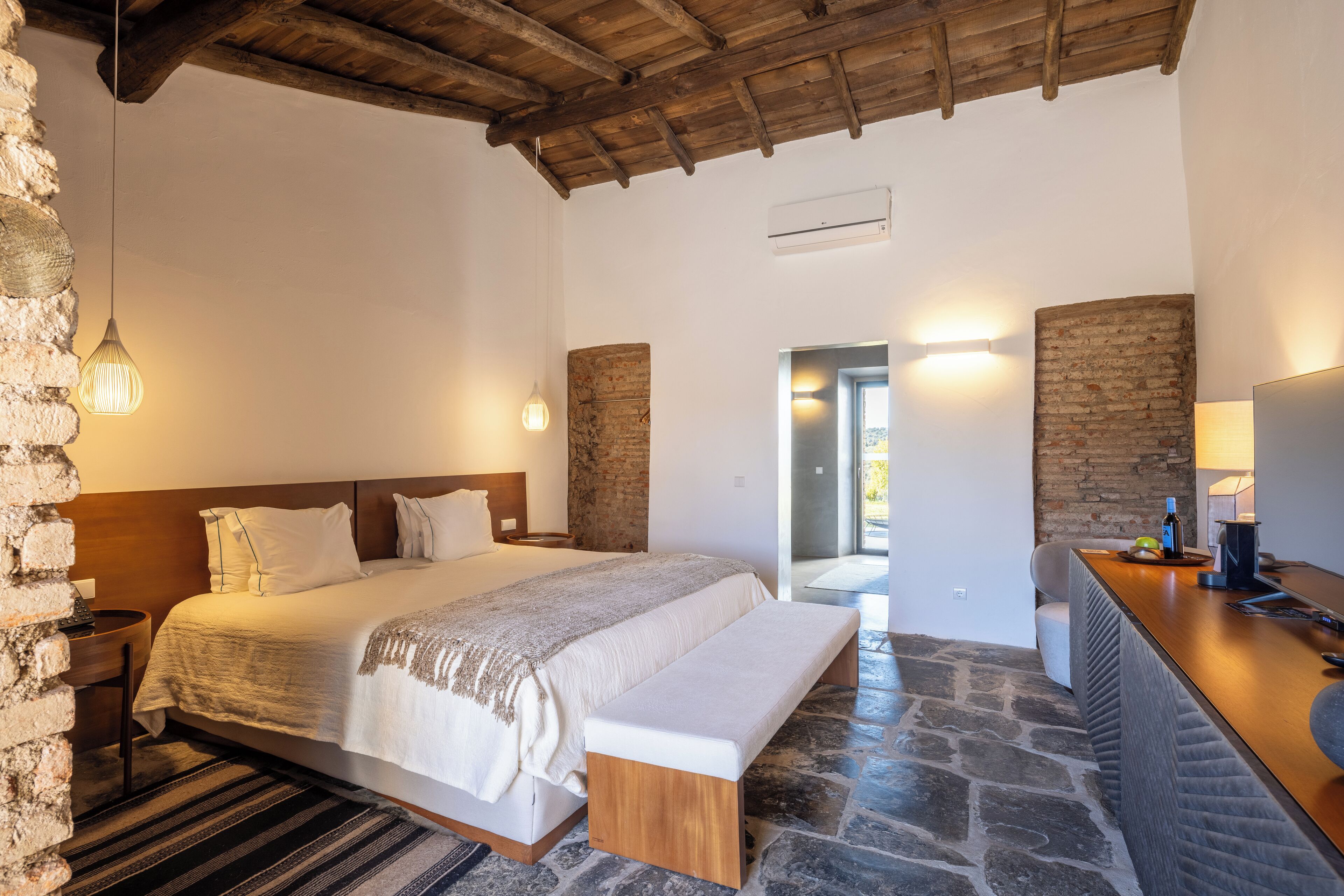 A bedroom with a large bed, a bench, a stone floor, a wooden ceiling, and a stone wall.