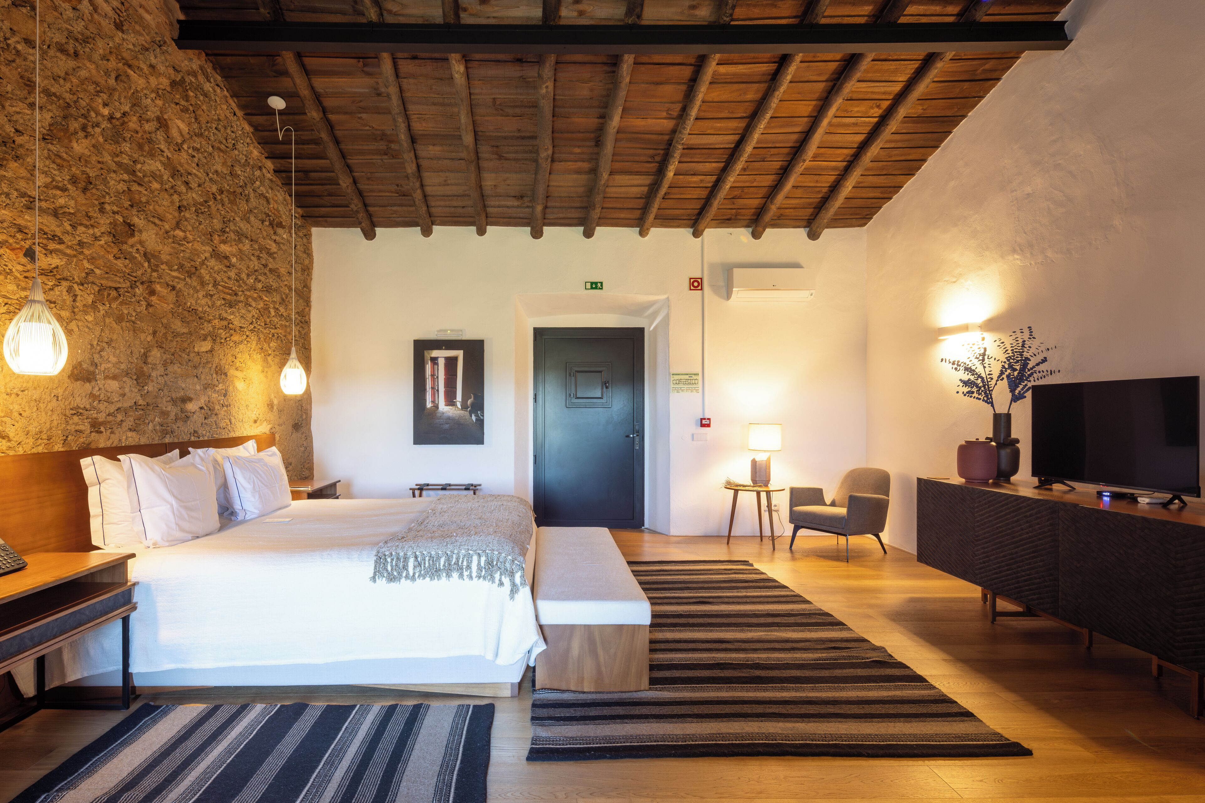 A hotel room with a wooden ceiling, a large bed, a desk, a TV, and a small table with a lamp.