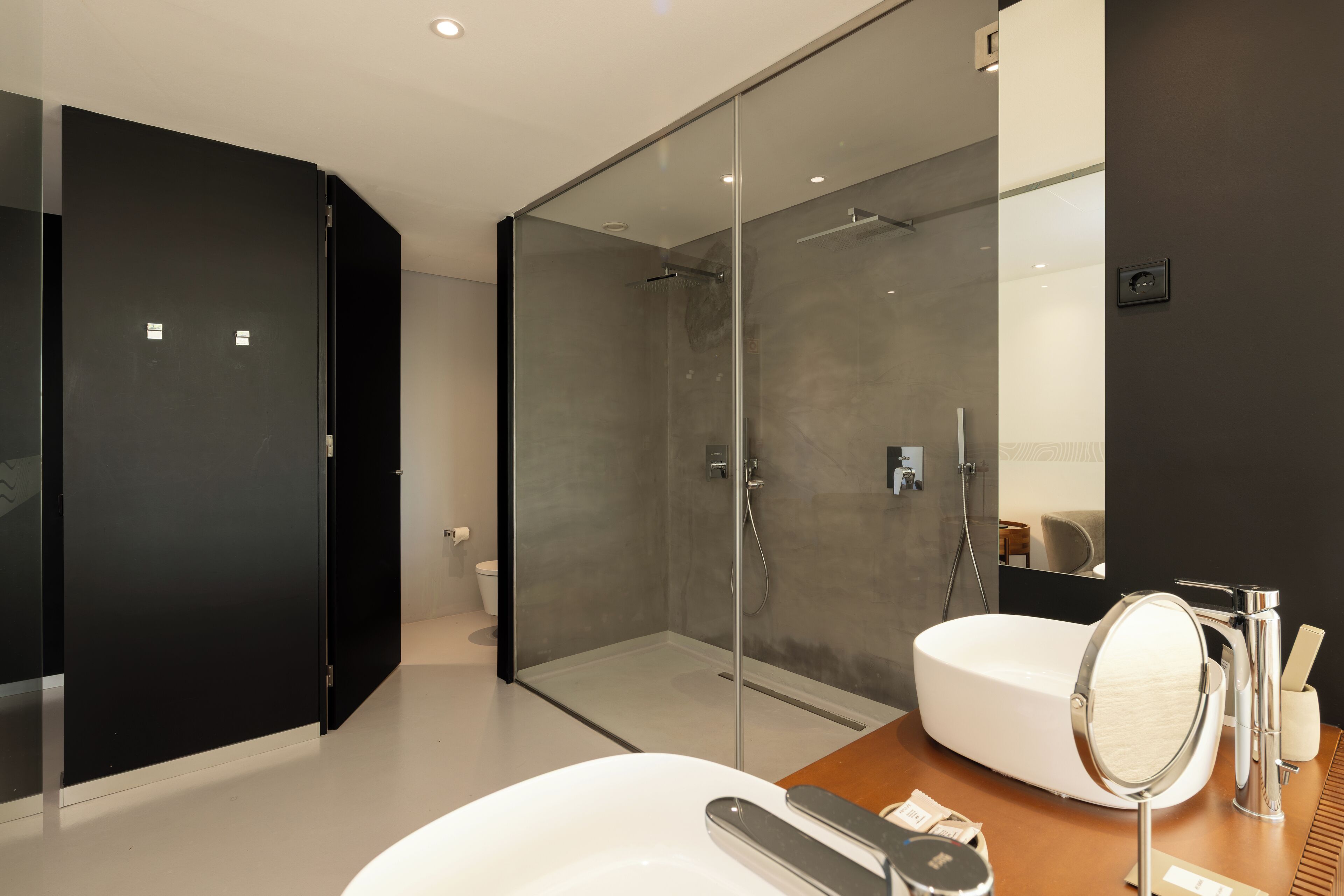 Un baño moderno con cabina de ducha de vidrio, un lavabo blanco con espejo redondo y un mueble de tocador de madera.