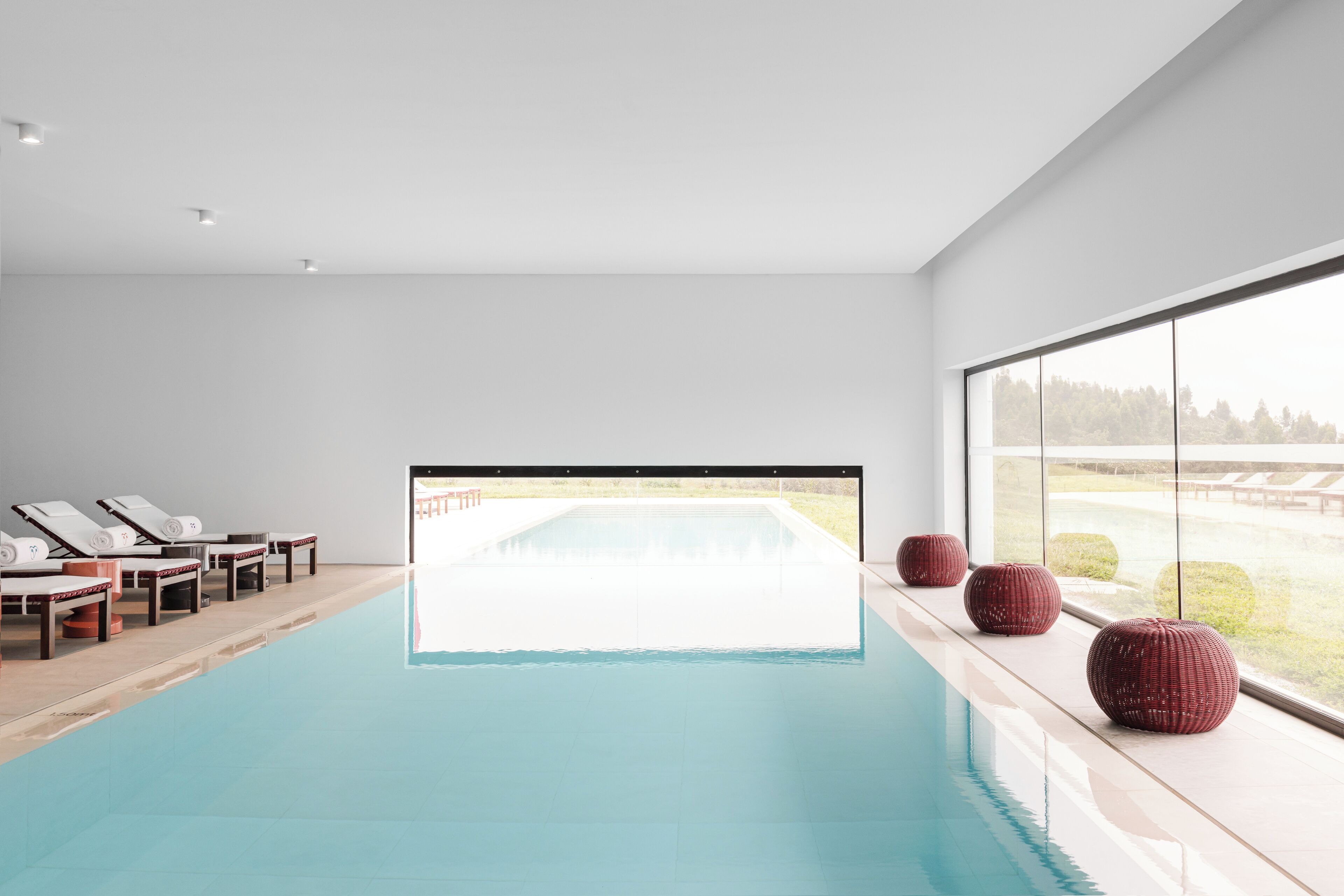 Una piscina interior moderna con paredes de vidrio transparente, sillas de descanso y un amplio ventanal con vistas al exterior.