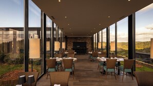 Un amplio comedor al aire libre con techos altos y ventanas grandes que ofrecen vistas del paisaje circundante. Las mesas están dispuestas con sillas y vajilla, y se observa una chimenea al fondo.