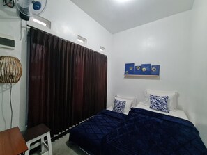 A bedroom with a bed, dark curtains, a small table, a wall-mounted fan, and a shelf with decorative items.