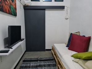 A small, compact room with a bed, a television on a stand, a desk with a keyboard, and a patterned rug.