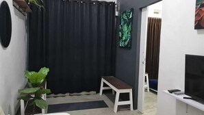 A compact living space with a dark curtain, a white bench, a small table, a TV, and a plant. - Omah Beji di Batu (Batu)