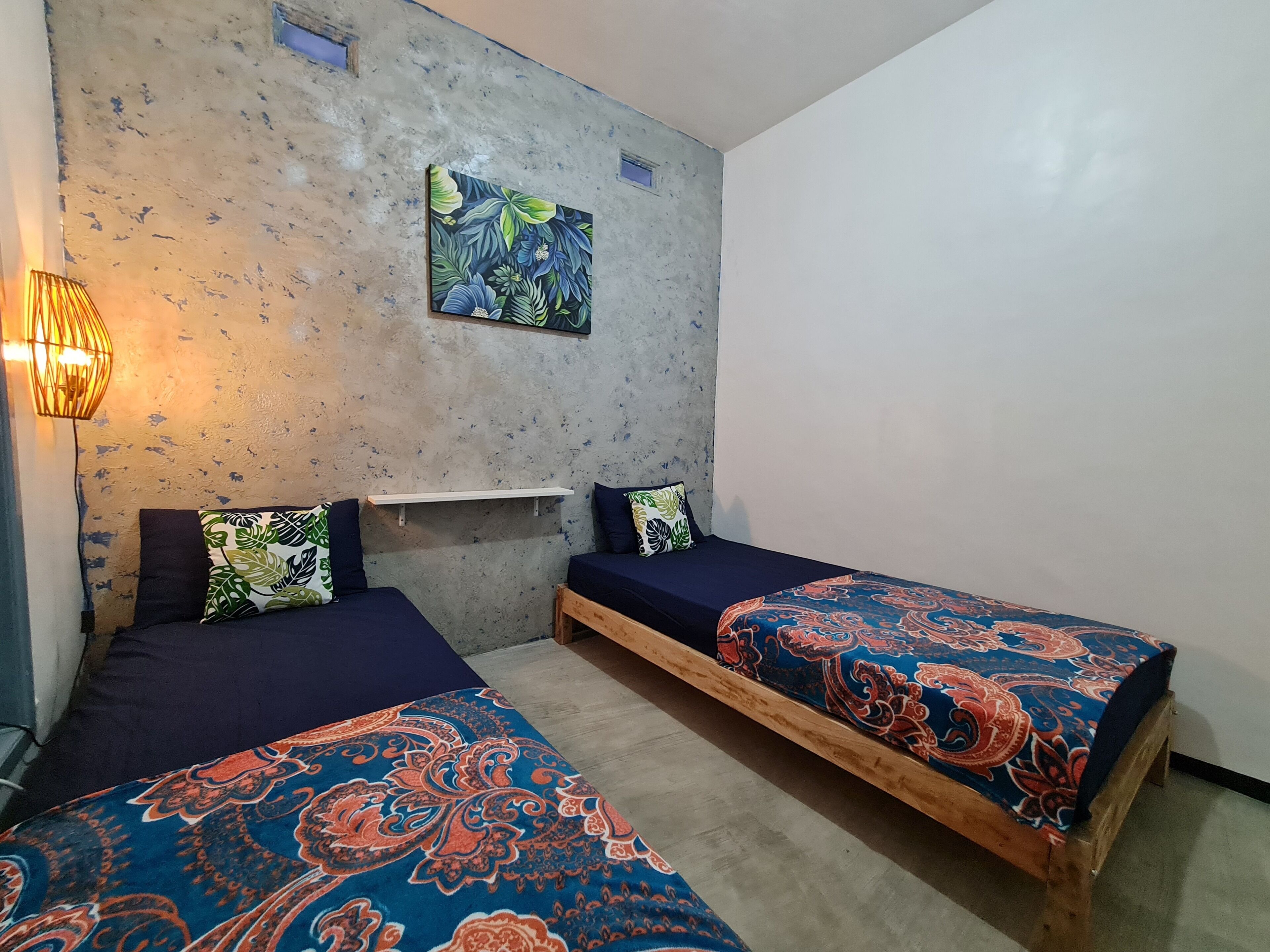 A room with two beds, a wall-mounted shelf, a painting, and a wall-mounted light fixture.