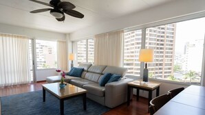 A modern hotel room with a sofa, coffee table, and lamp. There are large windows with curtains, a ceiling fan, and a view of tall buildings.