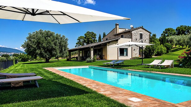 Une piscine bordée de pierres, entourée de pelouse, et une maison à la façade en pierre.