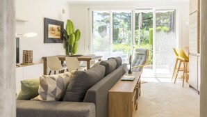 A modern living room with a sofa, a dining table, and chairs. There is a large window with a view of greenery outside. - Housing White Pearl Beach by Sharing Co (Marbella)