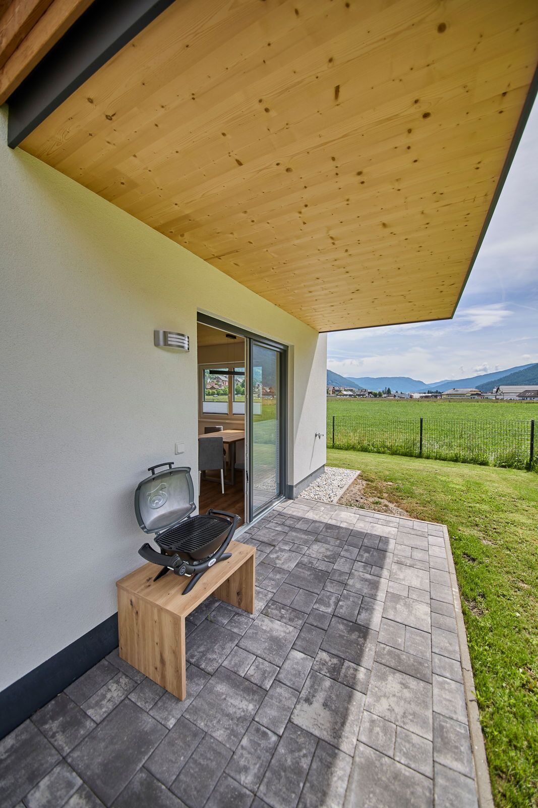 A patio with a grill, a wooden bench, and a view of a green field and mountains.