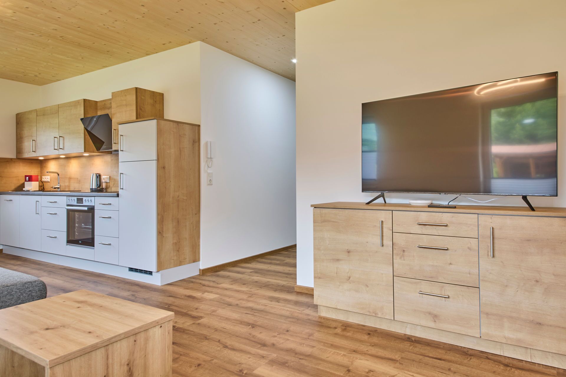 A modern kitchen with wooden cabinets, a built-in oven, and a flat-screen TV on a wooden stand.