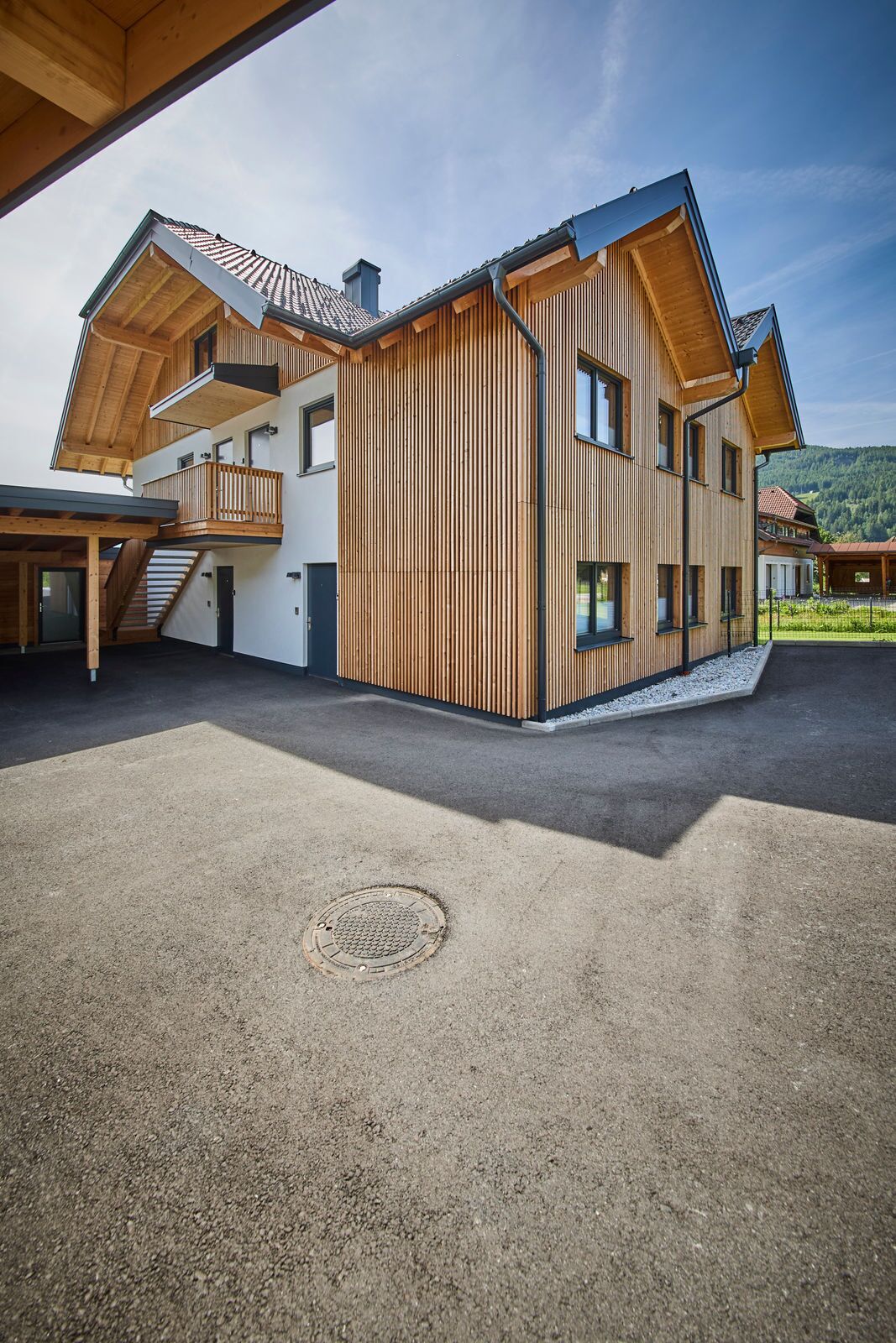 Un edificio moderno de varios pisos con paneles exteriores de madera, un acceso cubierto y un cielo despejado.