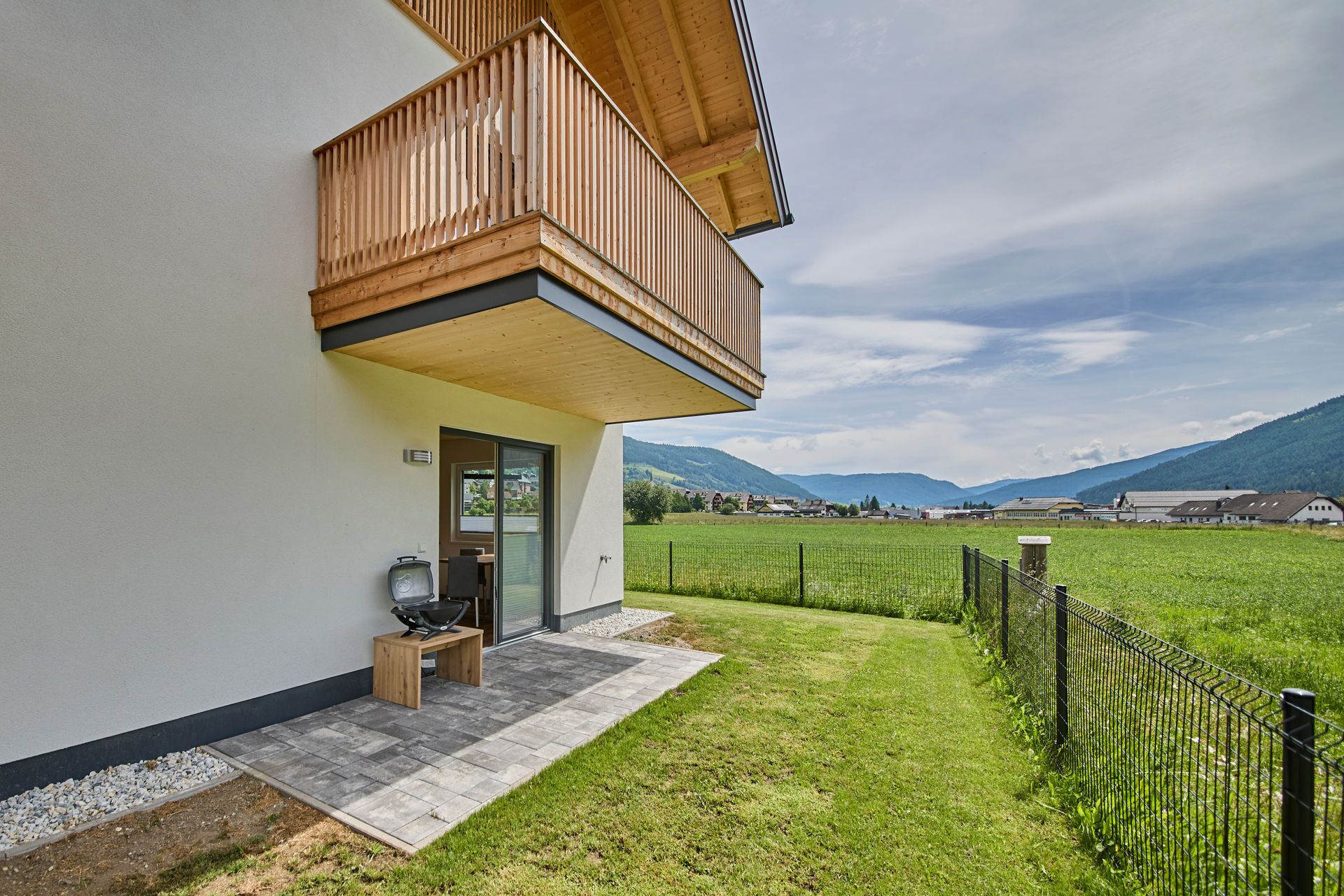 A modern building with a wooden balcony, a small outdoor seating area, and a view of a green field and mountains in the background.