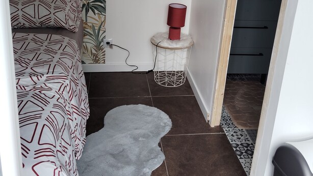 Une chambre avec une couvre-lit à motifs, un petit tapis, deux miroirs muraux, une lampe rouge et une poubelle blanche.