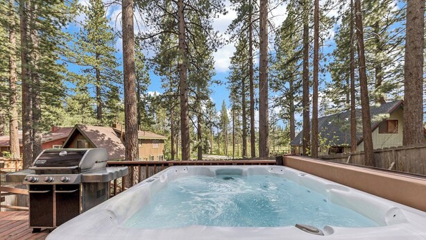 A hot tub on a wooden deck with a grill and a view of tall pine trees.