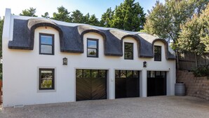 A modern house with a unique curved roof, multiple garage doors, and a gravel driveway. - Thatched Apartment with great views, full kitchen, Wifi in Noordhoek, Cape Town (Cape Town)