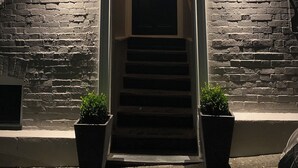 A building entrance with a dark brick facade, a lit sign above the door, and two potted plants at the bottom. - No22 (Barnstaple)