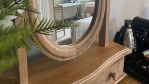 A wooden vanity with a mirror reflecting a room with a dining table, a chair, and a hanging light fixture. - No22 (Barnstaple)