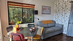 Una sala de estar con un sofá gris, una mesa de comedor con sillas, un ventanal con persianas, un mapa del mundo en la pared y una cortina con estampado de pájaros.