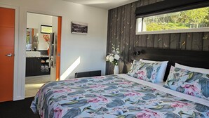 Un dormitorio con cama, ventilador de techo, ventana con vista a follaje, baño con espejo y puerta.