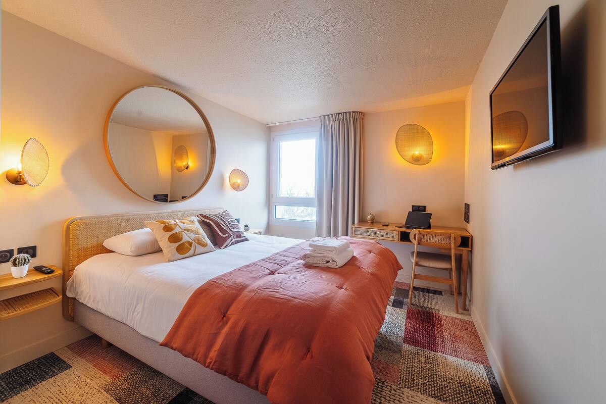 A hotel room with a large bed, a round mirror, a desk, and a window with curtains.