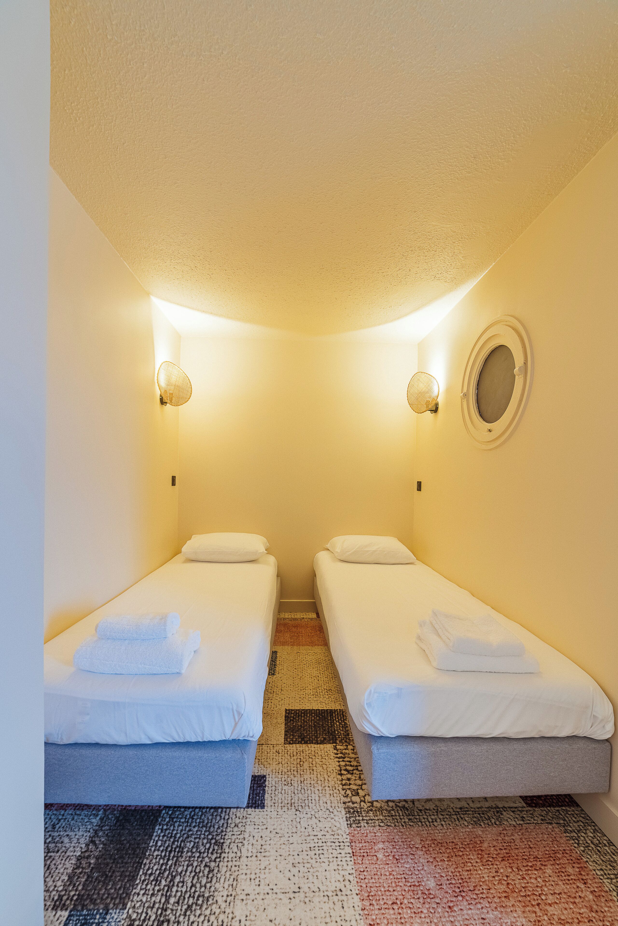 A small, well-lit room with two single beds, each with a pillow and a folded towel. There is a round mirror on the wall and a textured rug on the floor.