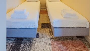 A small, well-lit room with two single beds, each with a pillow and a folded towel. There is a round mirror on the wall and a textured rug on the floor.