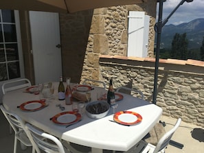 A table set for dining with wine bottles, glasses, and plates, set against a stone wall with a mountain view.