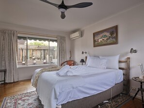 A bedroom with a bed, a window with curtains, a ceiling fan, an air conditioning unit, a painting on the wall, and a small table with a lamp.