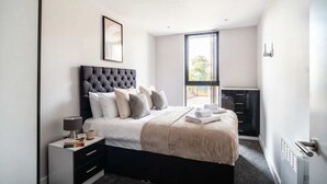 Un dormitorio moderno con un cabecero capitonado, una mesita de noche con lámpara, una cama bien hecha y vistas al exterior a través de un ventanal grande.