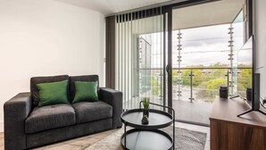 Una sala moderna con un sofá gris, una mesita redonda con una planta y un gran ventanal corredero que da a un balcón con vistas a la ciudad.