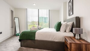 Un dormitorio moderno con una cama grande, una mesita de noche con lámpara, un espejo y vistas a edificios a través de amplias ventanas.