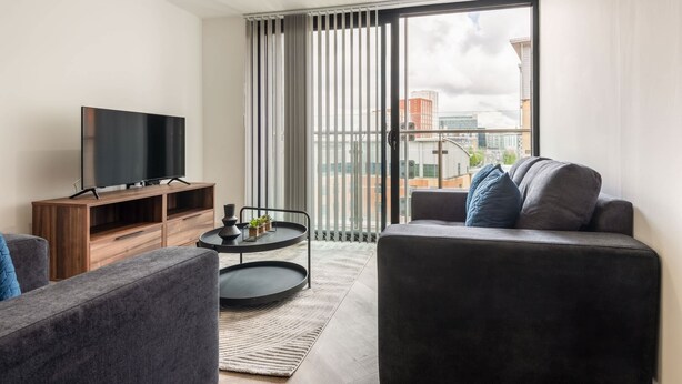 Ein modernes Wohnzimmer mit einem Flachbildfernseher auf einem Holzständer, einem grauen Sofa, einem kleinen runden Couchtisch mit Tablett und einer großen Schiebeglasdoor, die zu einem Balkon mit Stadtblick führt.