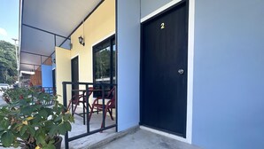 Un bâtiment avec une porte noire portant le numéro 2, un espace extérieur abrité avec des chaises rouges, et un pot de fleurs contenant une plante à feuilles vertes.