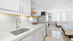 A modern kitchen with white cabinets, a stainless steel sink, and a built-in oven. There is a small dining area with a bench and a window providing natural light. - One Step to The Beach (Sunny Isles)