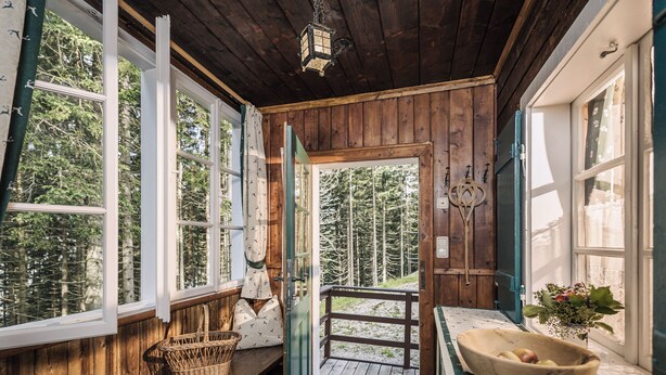 Una cabaña de madera con ventanas amplias, una banca, una cesta de mimbre, un cuenco de madera con manzanas y una puerta que da al exterior.