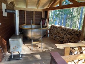 A sauna room with a large wooden tub, a metal heater, and a stack of firewood.