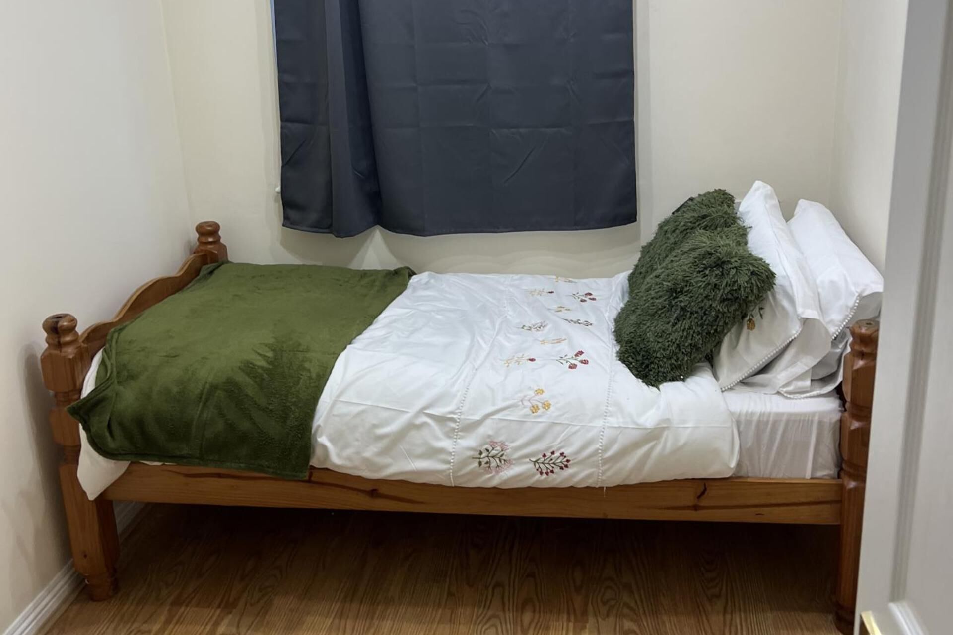 A small, single-bed room with a wooden frame, a green blanket, white bedding with floral embroidery, a green pillow, and a dark blue curtain.