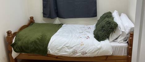 A small, single-bed room with a wooden frame, a green blanket, white bedding with floral embroidery, a green pillow, and a dark blue curtain.