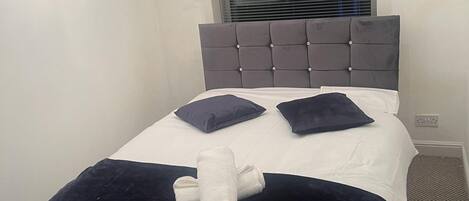 Una cama bien hecha con un cabecero gris, dos almohadas y una toalla enrollada.