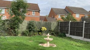 A garden with a central fountain, surrounded by a wooden fence and residential buildings in the background. - Cozy 3 Bedrooms Birmingham House With Pool Table (Birmingham)