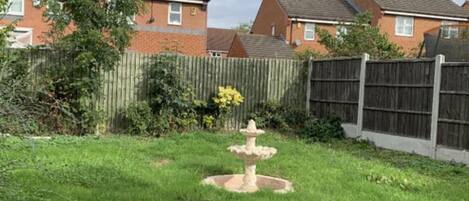 Un jardín con una fuente central, rodeado por una valla de madera y edificios residenciales al fondo.