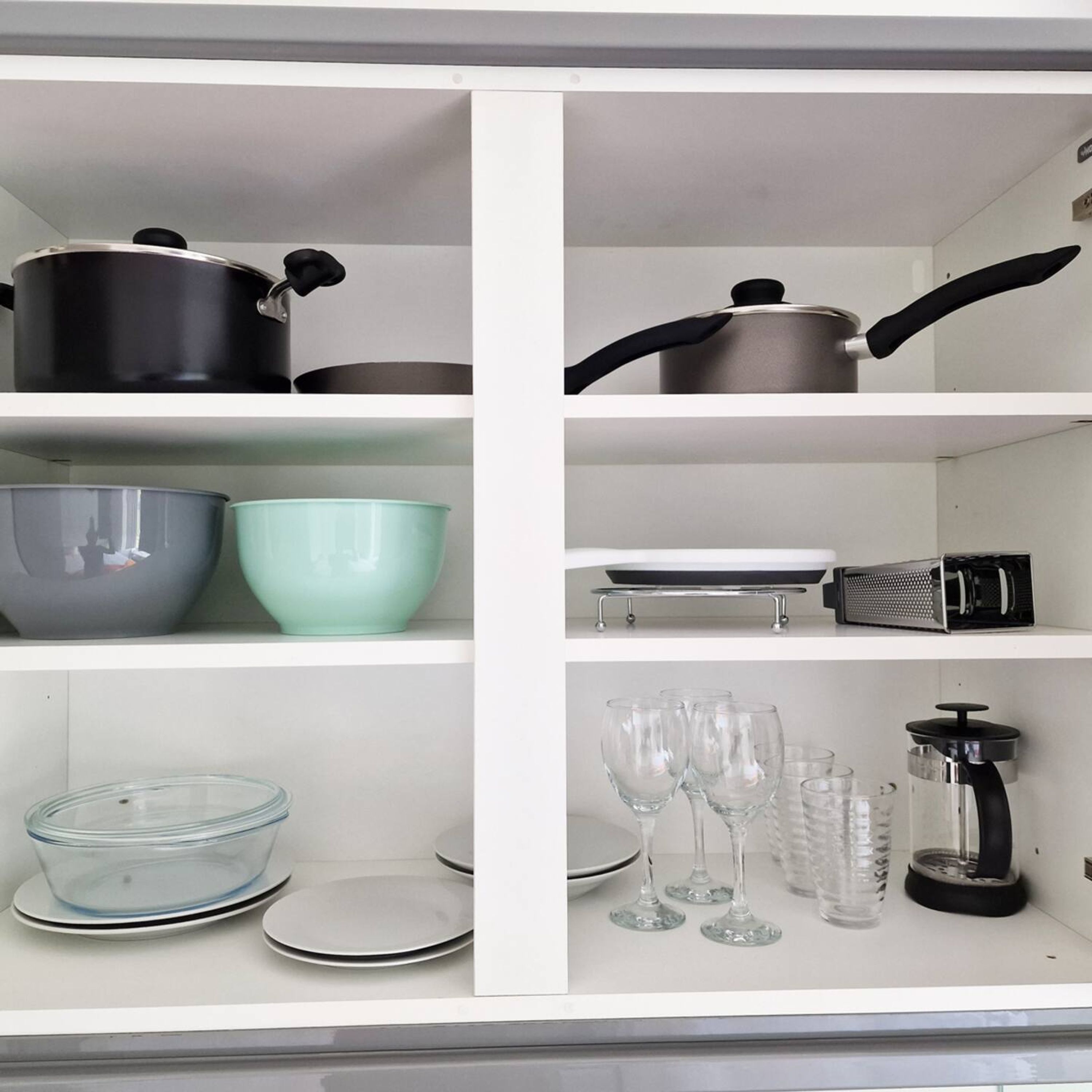 A kitchen with white cabinets, black and grey pots, a green bowl, a grater, and clear glassware.