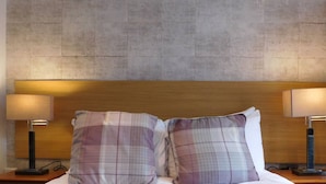 Una cama bien hecha con un cojín escocés y una manta con texto, flanqueada por dos lámparas de noche sobre mesitas de madera.
