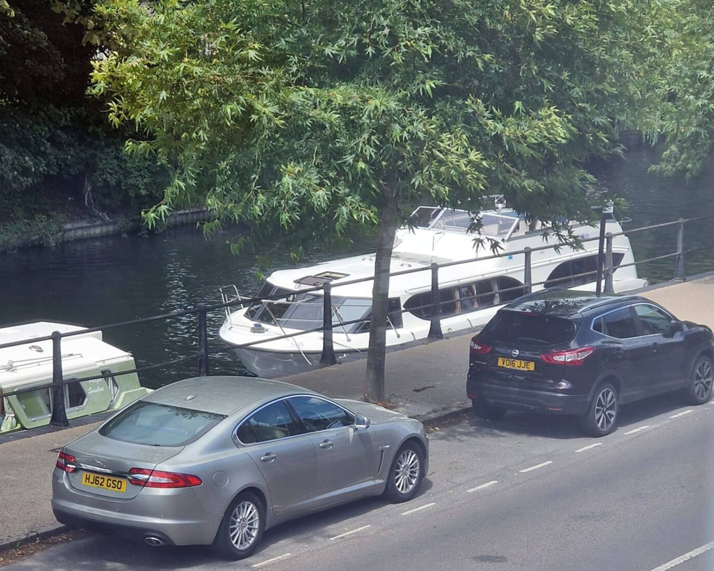 A river with boats docked alongside a road with parked cars.