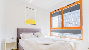 Une chambre à coucher comprenant un lit, une table de chevet, une fenêtre donnant sur des immeubles et une œuvre d’art accrochée au mur.