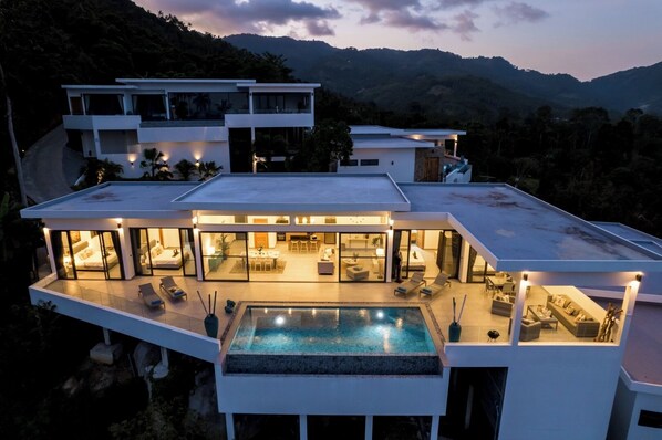 A modern multi-story house with a rooftop pool, large glass windows, and a view of the surrounding hills.