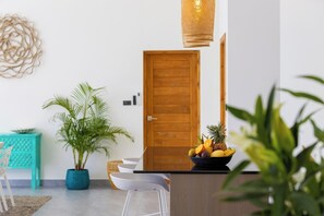 A modern kitchen with a wooden door, a hanging light fixture, a fruit bowl on the counter, and a potted plant.