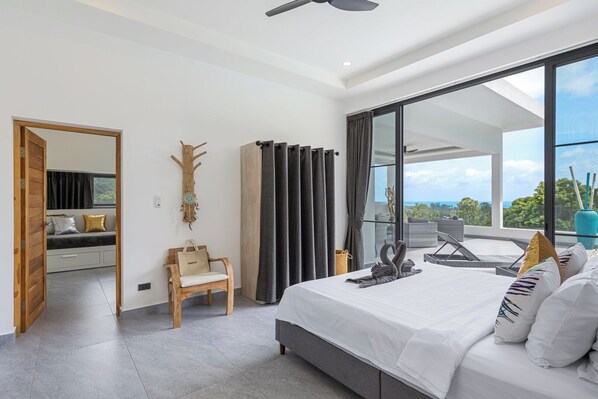 A modern bedroom with a large bed, a seating area with a sofa and chair, a decorative wall piece, and a balcony with a view.
