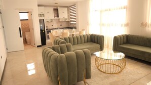 A modern living room with a green sofa set, a glass coffee table, and a kitchen area with white cabinets and a black countertop. - Yohannes Guesthouse Addis (Addis Ababa)