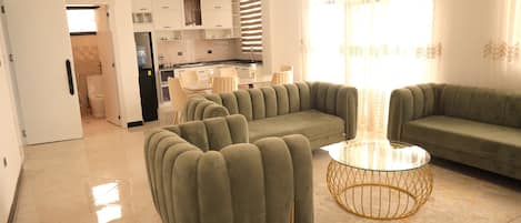 Un salon moderne avec un canapé vert, une table basse en verre et un coin cuisine doté de meubles blancs et d’un plan de travail noir.